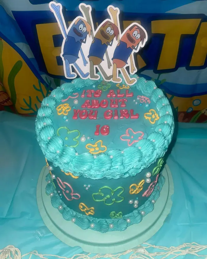 A tall teal buttercream cake with shell piping, pearls and colourful piped flowers, reading It’s all about you girl 16